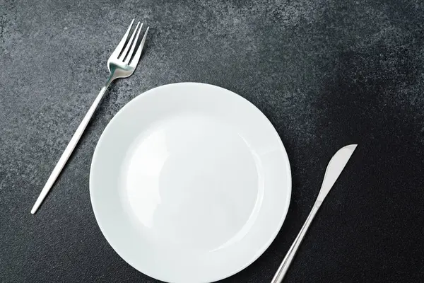 Temiz, basit bir tablo görüntüsünde yiyecek bekleyen boş bir tabak ve çatal bıçak bulunur..