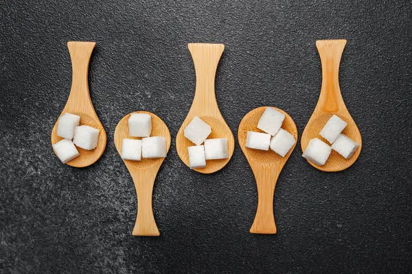 Tahta kaşıklar düzgün yerleştirilmiş şeker küplerini karanlık bir taş zemin üzerinde tutarlar..