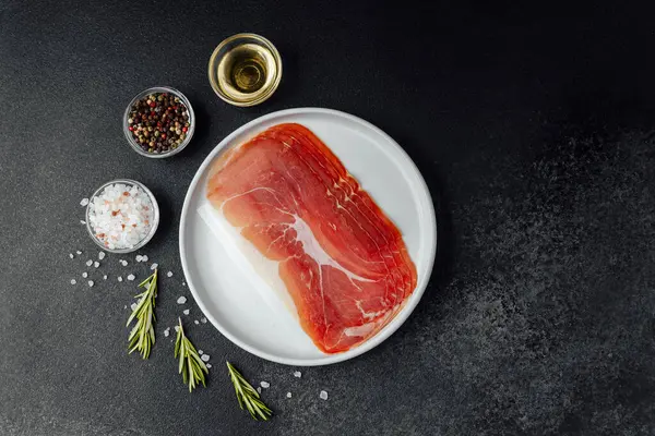 Beyaz tabakta dilimlenmiş jambon ve siyah arka planda baharatlar.