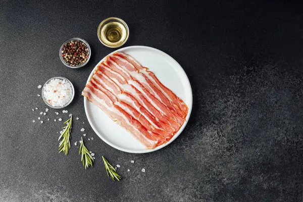 Beyaz tabakta çiğ domuz pastırması, biber, tuz ve biberiye.