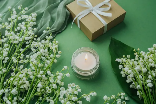 Vadideki zambaklarla çevrili yanan mum ve yeşil arka planda paketlenmiş hediye.