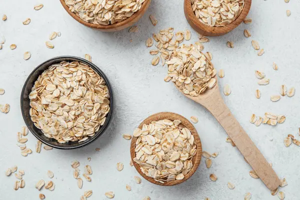 Çiğ yulaf dolu kaseler yakınlarda tahta bir kaşıkla birlikte bir yüzeye oturur..