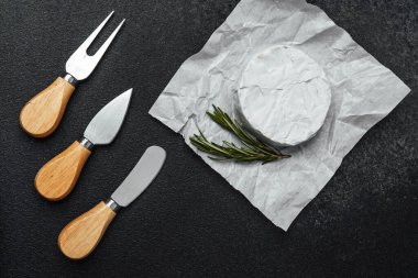 Peynir aletleri ve taze otlar buruşmuş kağıtta yuvarlak bir peynirin yanında..