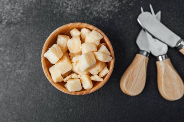 Taze peynir küpleriyle dolu ahşap bir kase, karanlık bir yüzeyde peynir aletlerinin yanında oturur..