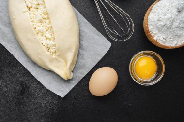 Süzme peynirli çiğ khachapuri ve siyah arka plandaki malzemeler.