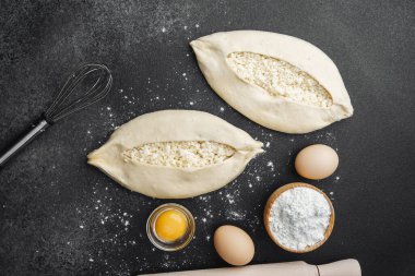 İki çiğ khachapuri ekmeği, içinde fırın malzemeleri olan siyah un kaplı bir masada.