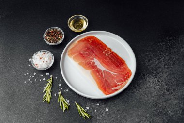 Beyaz tabakta dilimlenmiş jambon ve siyah arka planda baharatlar.
