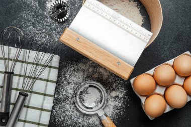 Yumurtalar, un ve çeşitli pişirme aletleri karanlık bir mutfak tezgahında düzenlenir..