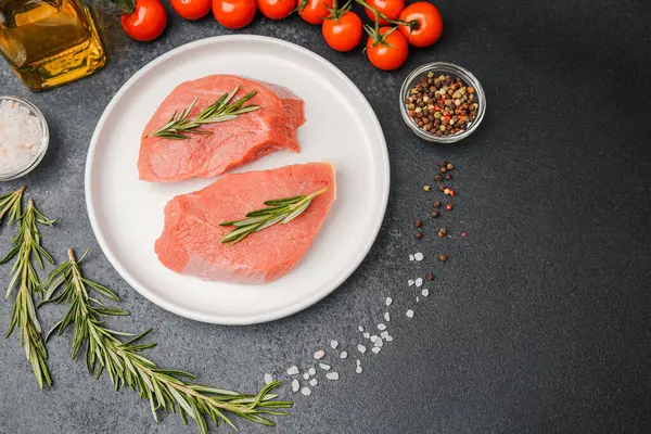 Beyaz seramik tabakta çiğ biftek, domates, tuz ve biber.
