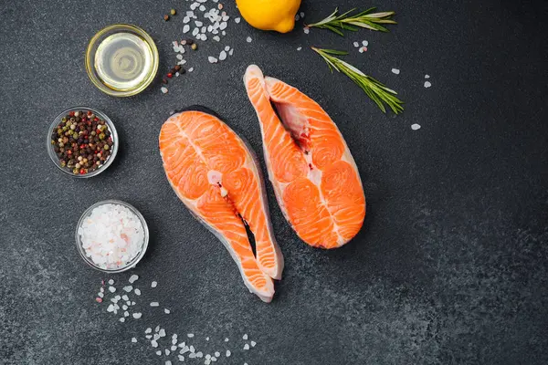 Somon biftekleri siyah bir tezgahta otlar, limonlar ve baharatlarla sıralanır..