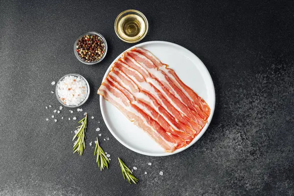 Beyaz tabakta çiğ domuz pastırması, biber, tuz ve biberiye.