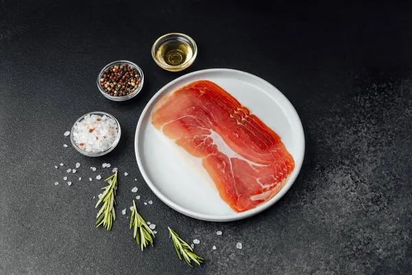 Beyaz tabakta dilimlenmiş jambon ve siyah arka planda baharatlar.