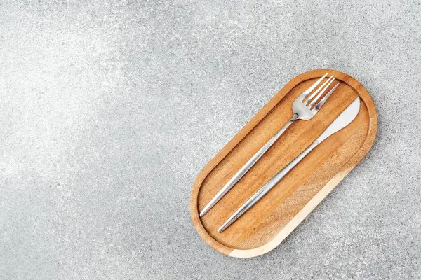 Ahşap bir tabak, gri arka planda düzgün yerleştirilmiş çatal ve bıçak taşır..