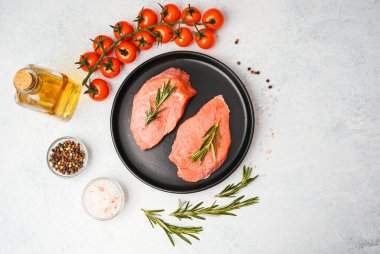 Siyah tabakta biberiyeli iki çiğ biftek ve beyaz yüzeyde pişirme malzemeleri.