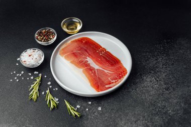 Beyaz tabakta dilimlenmiş jambon ve siyah arka planda baharatlar.