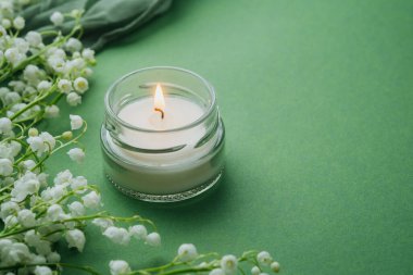 Yeşil arka planda beyaz zambaklı cam mum yanıyor.