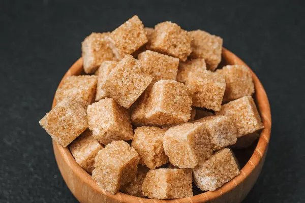 Esmer şeker küpleri siyah bir arka plana karşı ahşap bir kaseyi doldurur..