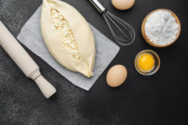 Süzme peynirli çiğ khachapuri ve siyah arka plandaki malzemeler.