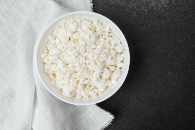 Bir kase yumuşak süzme peynir, yakınlarda beyaz bir bezle koyu bir tezgahın üzerinde duruyor..