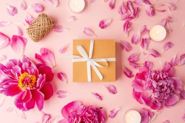 Beyaz kurdeleli hediye kutusu şakayıklar, taç yaprakları, mumlar ve pembe arka planda sicimle çevrili.