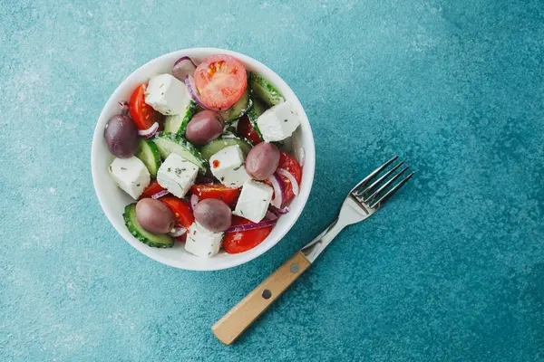 Beyaz kâsede beyaz peynirli Yunan salatası ve mavi arka planda çatal.