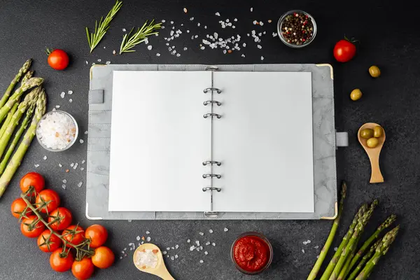 Koyu arkaplanda yemek malzemeleriyle çevrili boş sayfaları olan tarif defteri