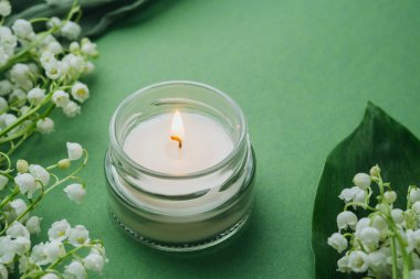 Yeşil arka planda beyaz zambaklı cam mum yanıyor.