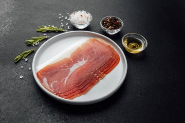Beyaz tabakta dilimlenmiş jambon ve siyah arka planda baharatlar.