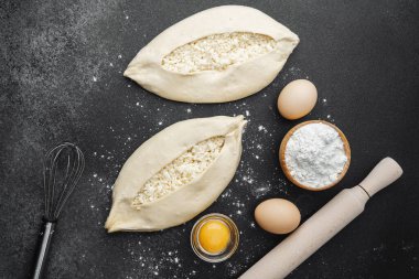 İki çiğ khachapuri ekmeği, içinde fırın malzemeleri olan siyah un kaplı bir masada.