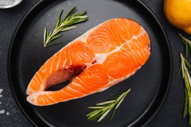 Canlı bir somon bifteği siyah bir tabakta otlar ve limon dilimleriyle sunulur..