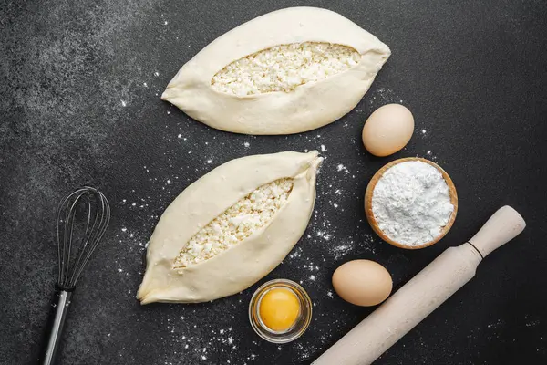 İki çiğ khachapuri ekmeği, içinde fırın malzemeleri olan siyah un kaplı bir masada.