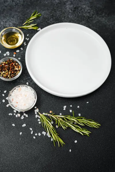 Siyah taş zemin üzerinde bitkiler ve baharatlar olan boş beyaz tabak