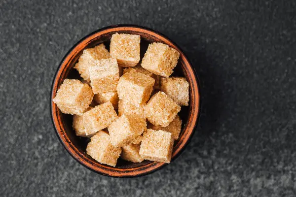 Kahverengi şeker küpleriyle dolu bir kase koyu renkli bir taş yüzeyde duruyor..