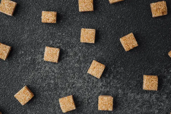Küçük kahverengi şeker küpleri koyu bir yüzey boyunca dağılır ve desenli bir görünüm oluşturur..