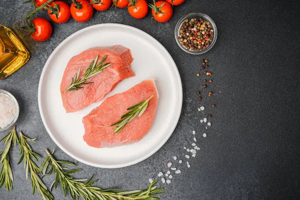 Beyaz seramik tabakta çiğ biftek, domates, tuz ve biber.