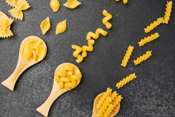 Koyu bir tezgahın üzerinde ahşap kaşıklarla hazırlanmış farklı şekillerde pişmemiş makarna..