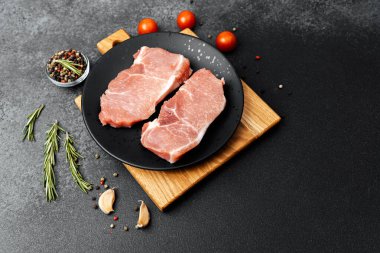 Mutfakta baharat ve otlarla çevrili siyah bir tabakta iki çiğ domuz pirzolası..