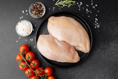 Siyah tabakta çiğ tavuk göğsü, domates ve koyu arkaplanda baharat.