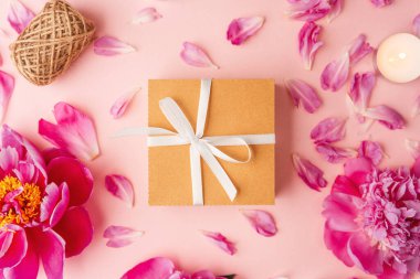 Beyaz kurdeleli hediye kutusu şakayıklar, taç yaprakları, mumlar ve pembe arka planda sicimle çevrili.