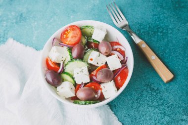 Beyaz kâsede beyaz peynirli Yunan salatası ve mavi arka planda çatal.