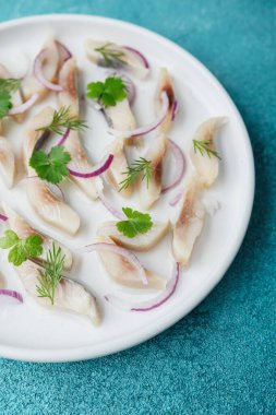 Kırmızı soğanlı marine edilmiş ringa dilimleri ve mavi arka planda beyaz tabakta otlar.