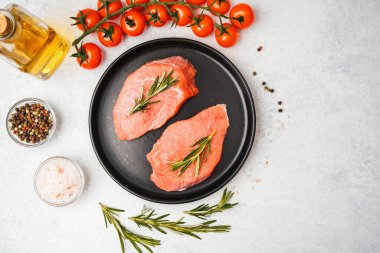 Siyah tabakta biberiyeli iki çiğ biftek ve beyaz yüzeyde pişirme malzemeleri.