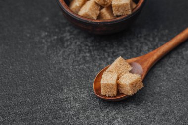 Kahverengi şeker küpleri ahşap bir kaşığın içine, desenli bir yüzey üzerinde küçük bir kasenin yanına yerleştirilmiş..