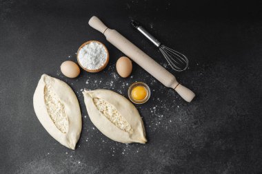 İki çiğ khachapuri ekmeği, içinde fırın malzemeleri olan siyah un kaplı bir masada.