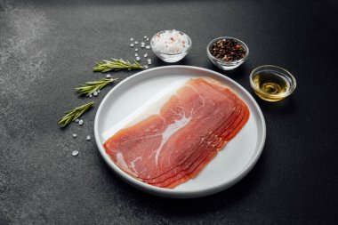 Beyaz tabakta dilimlenmiş jambon ve siyah arka planda baharatlar.