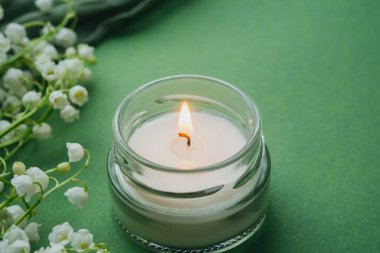 Yeşil arka planda beyaz zambaklı cam mum yanıyor.