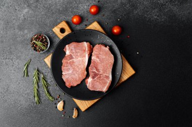 İki çiğ domuz pirzolası taze otlar ve domateslerle çevrili siyah bir tabağa konur..