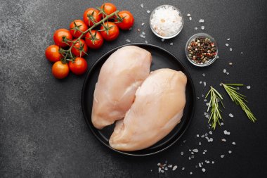 Siyah tabakta çiğ tavuk göğsü, domates ve koyu arkaplanda baharat.