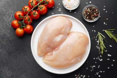 Beyaz tabakta çiğ tavuk filetosu, siyah masada tuz, domates ve otlar.