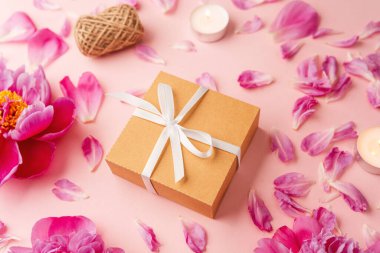 Beyaz kurdeleli hediye kutusu şakayıklar, taç yaprakları, mumlar ve pembe arka planda sicimle çevrili.